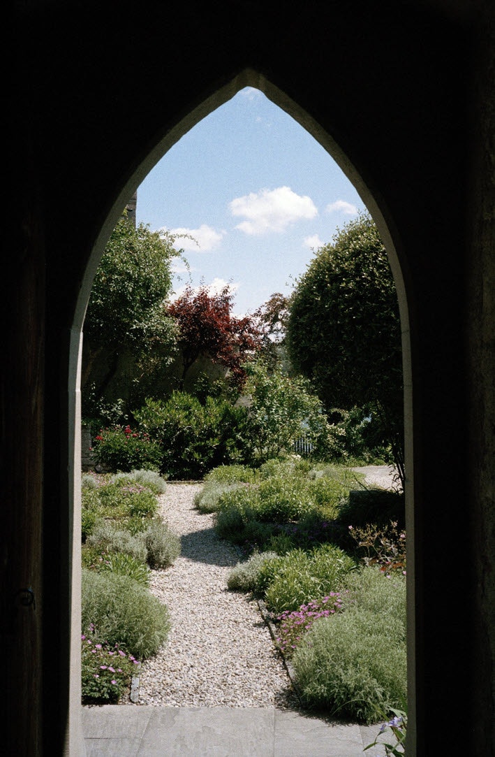 Blick durch eine Türe mit gotischem Spitzbogen ins Freie. Von der Schwelle der Türe führt Kiesweg in einen Garten. Zu beiden Seiten des Wegs wachsen Polsterpflanzen, einzelne von ihnen blühen in einem hellen Rosa. Im Hintergrund stehen Büsche. Über ihnen ist der Himmel zu sehen. Schönwetterwolken segeln über das Blau des Himmels.