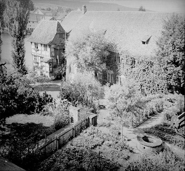 View from high-up of a garden and monastery buildings. There are plain tiles on the roofs. The walls are ivy-clad. Photo in black and white.